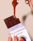 Chocolate bar with caramel being poured over it, held by a hand against a beige background.